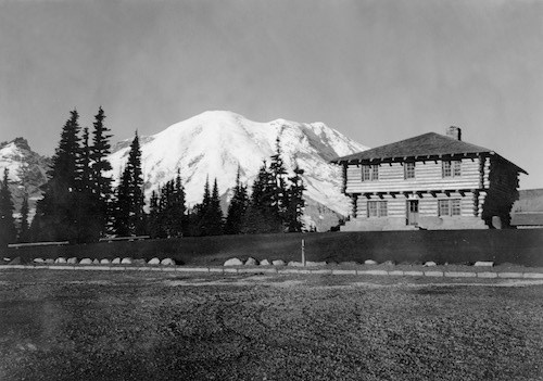 Black and white historic photo of a two-story square log building in front of a glaciated mountain peak.