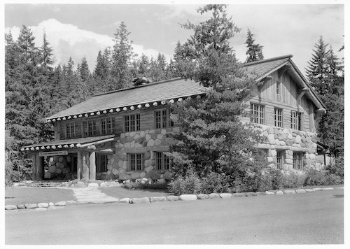 Black and white historic photo of a two-story building. The bottom story is clad in large boulders while the top story is built of logs with a wood shingle roof and covered porch.