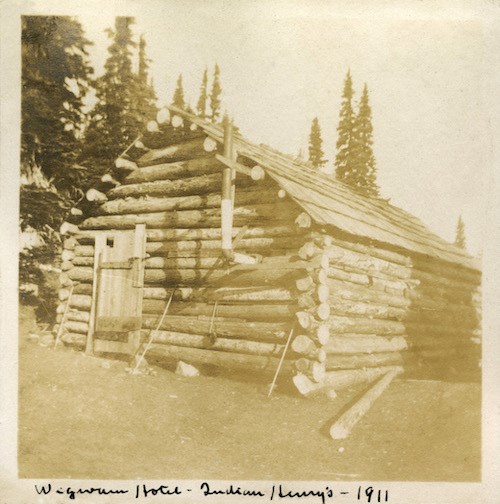 Historic photo of a roughly constructed log cabin with a single door, no windows and a metal smoke stack sticking out of the side of the cabin.
