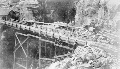Black and white photo of a stone bridge under construction framed by wood scaffolding with piles of debris on either side.