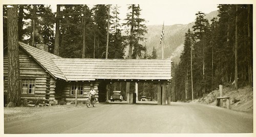 A log building with a roof extending out over two lanes of a dirt road. A person rides a bicycle past the building.