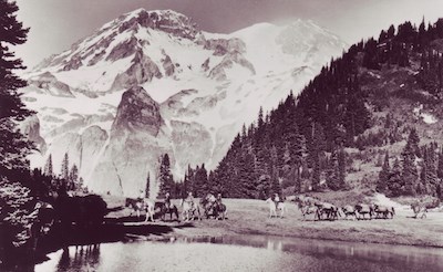 A string of horseback riders circle a subalpine lake with Mount Rainier rising in the background.