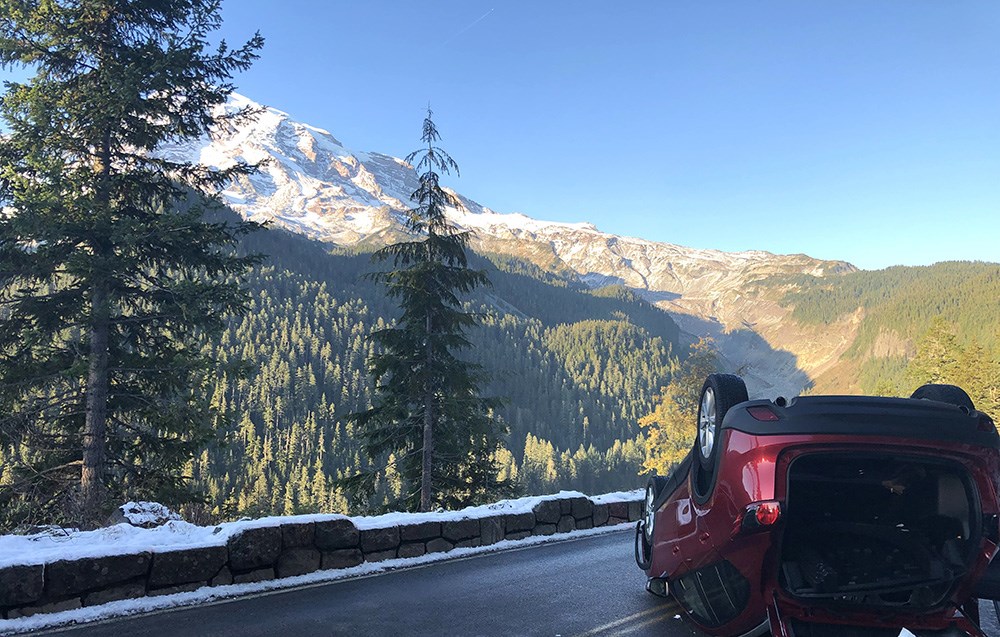 A red car flipped on its back along a steep, narrow road bordered by a snowy rock wall and overlooking a view of river valley and glaciated mountain.