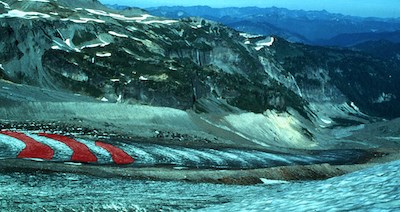 A glacier with bands of light and dark ice, with three dark bands highlighted in red, flows between rocky ridges.