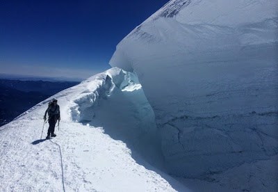 A climbing ranger stands at the edge of a large, deep crevasse on a steep slope.