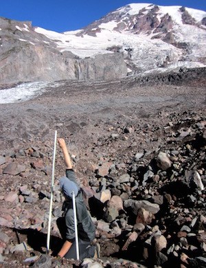 A person reaches up to measure the top of an ablation stake emerging from the rocky debris covered surface of a glacier that extends up towards the summit of Mount Rainier.