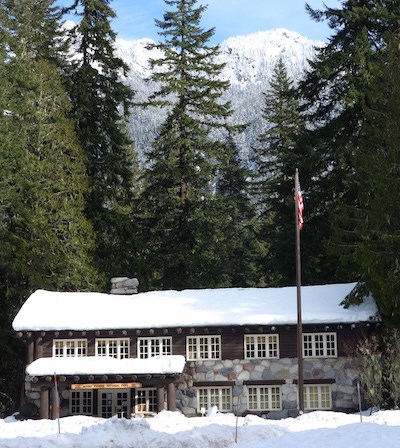 Rustic building with snow covered roof framed by conifer trees.