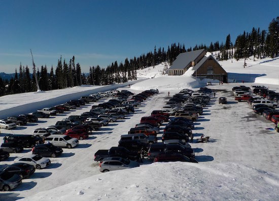 Cars fill a parking lot in front of a building with a steep roof and surrounded by deep snow banks on a sunny day.