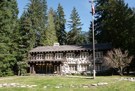 A historic wood and stone structure stands before tall evergreens and blue sky.