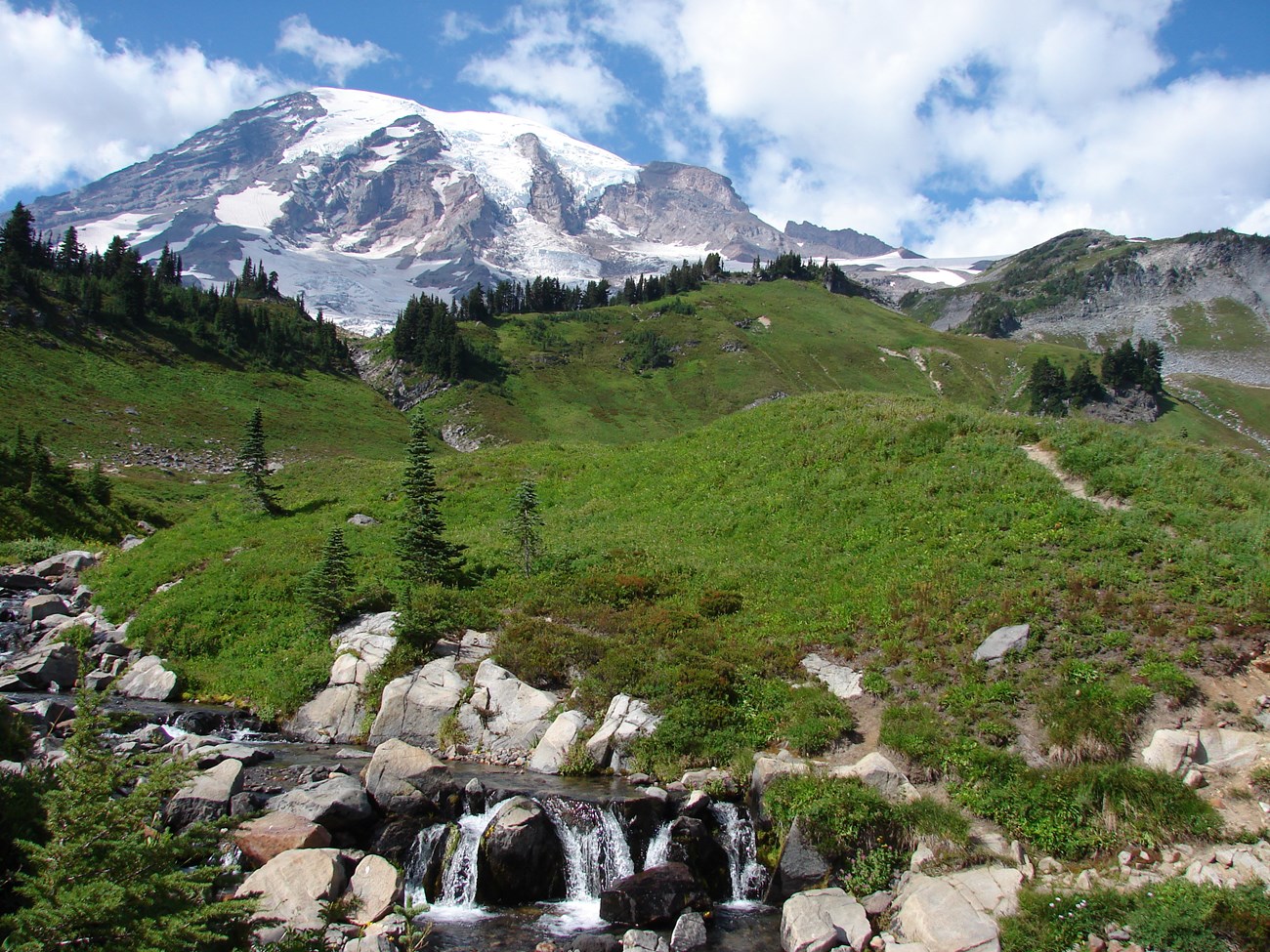 A cascading waterfall flows through the foreground. A lush green hillside and Mount Rainier fill the background.