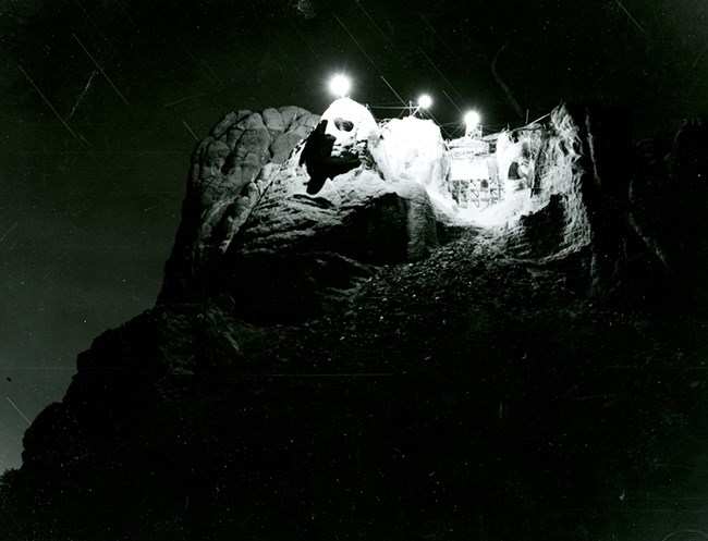 Black and white image of an attempt in 1939 to illuminate Mount Rushmore from the top of the sculpture.  Three lights shine from the top of the sculpture.  Scaffolding is visible in front of the Roosevelt face.