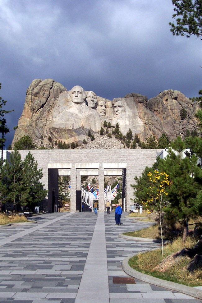 View of the walkway leading towards Mount Rushmore as it looks today.  The surface is made of different sized paving stones that are different shades of gray.  Mount Rushmore is in the background in the upper part of the image.