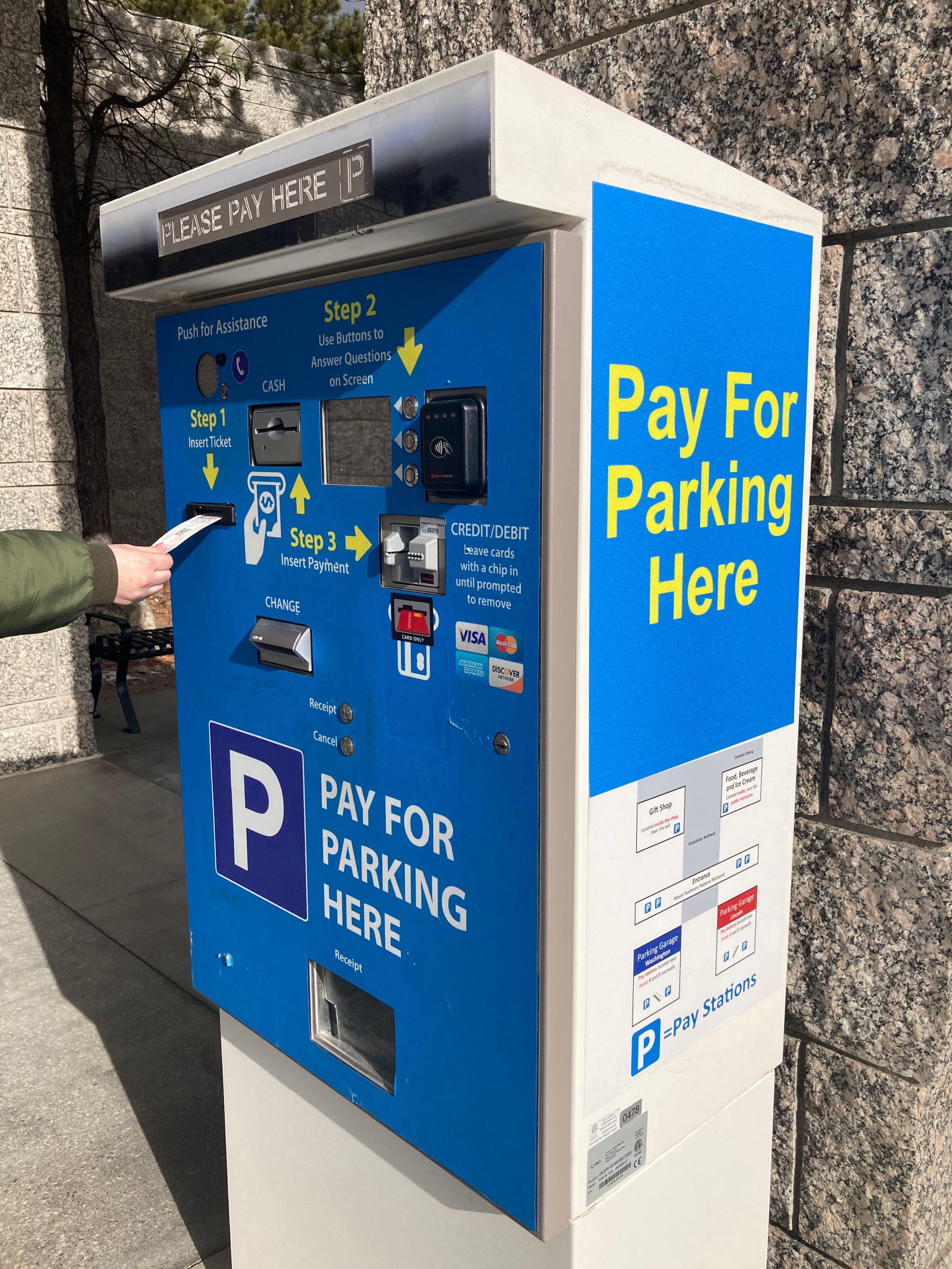 A visitor inserts a ticket into a pay-on-foot station to pay for parking. The station has blue panels and text in yellow letters instructing visitors to pay for parking here on the side and step-by-step instructions on the front.