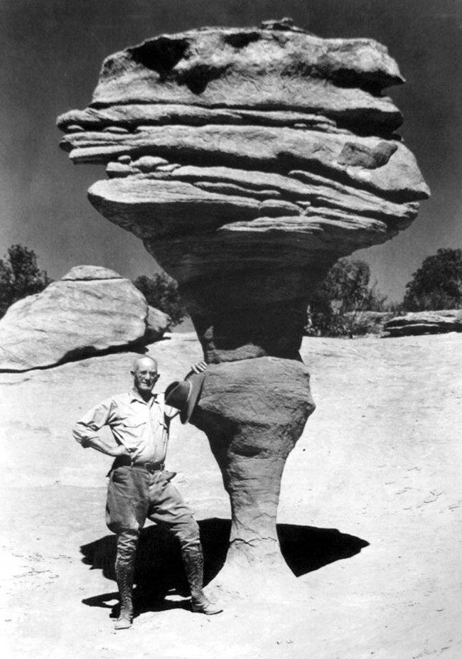 historical black and white photo of a man leaning against a large rock formation precariously perched on a spindly base