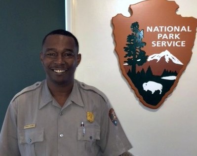 Smiling man wearing grey shirt with NPS badge over right pocket and NPS arrowhead on right sleeve