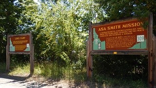 Two interpretive signs, one for Asa Smith Mission, and one for Long Camp stand on a highway pullout with trees in the background.