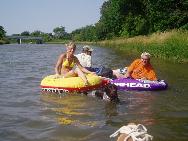 Three people and a dog enjoy the river on inflatable tubes