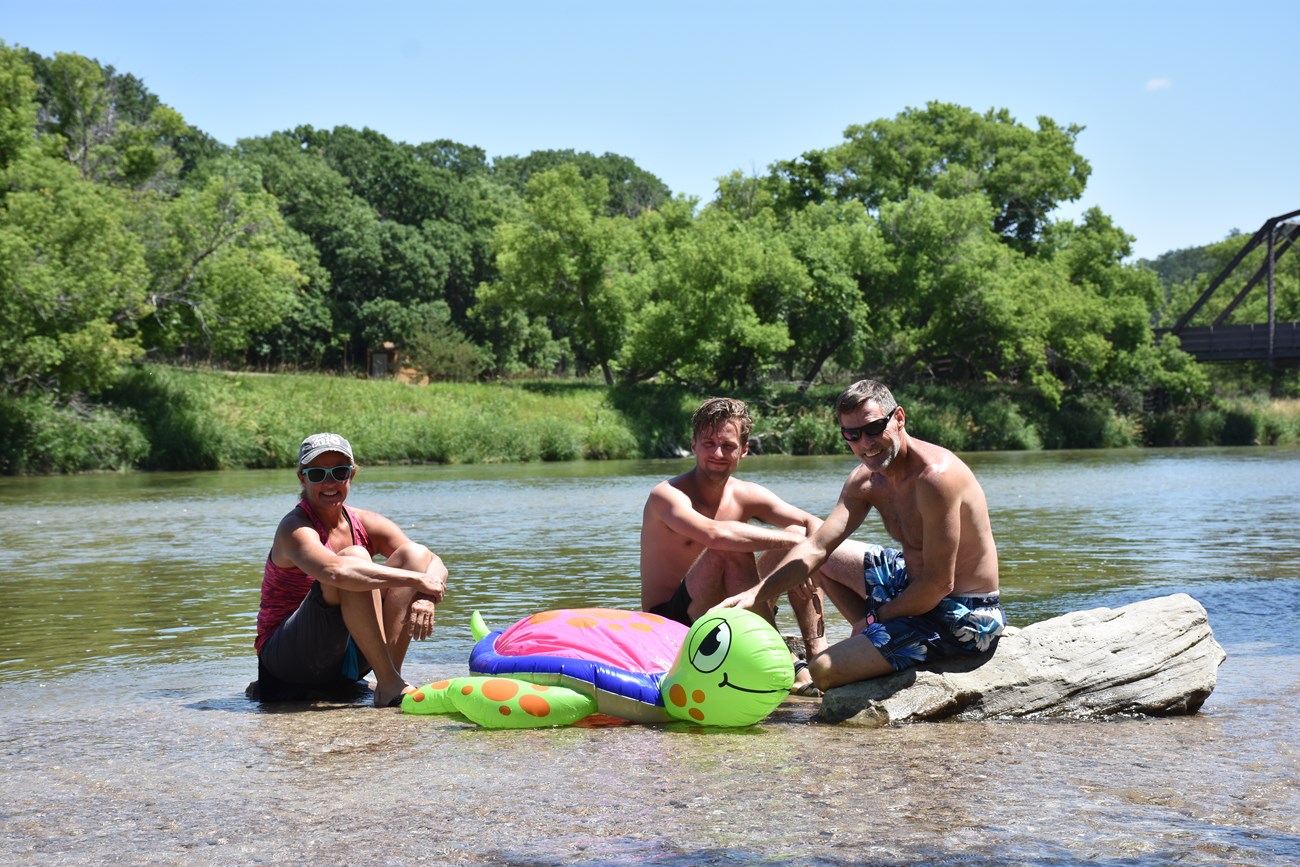 Three people setting in the shallows with an inflatable turtle.