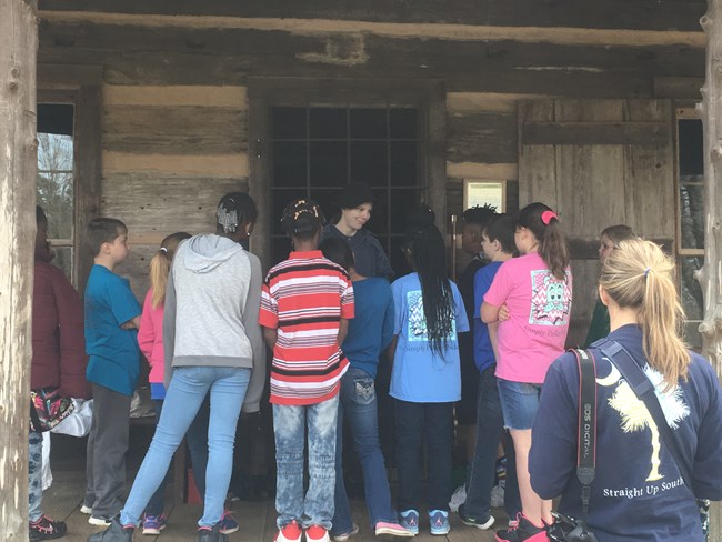 Children on a field trip gather around a reenactor at a log house.