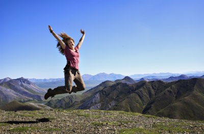 girl in pink shirt jumping on top of a mountain with more mountains in the distance