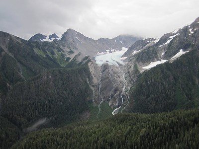 White Glacier recedes up the rocky Olympic mountains, with towering jagged peaks above and leaving waterfalls that drain into a lush green valley below.