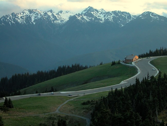 The Hurricane Ridge Visitor Center sits along a winding road, surrounded by grassy meadows with the Olympic Mountains in the distance.