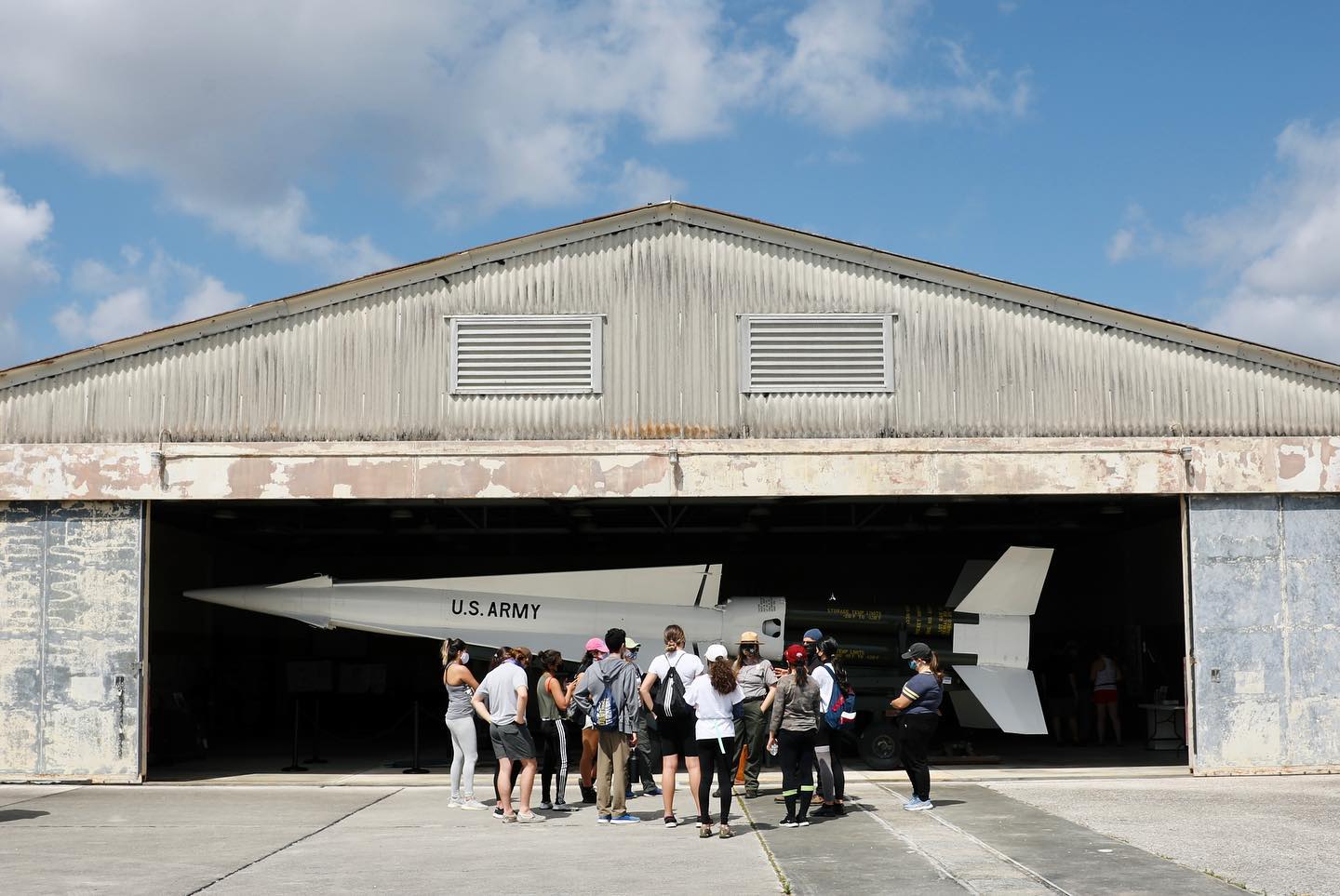 Several people face away, looking at a low hangar; inside is a white plane labeled "U.S. Army".