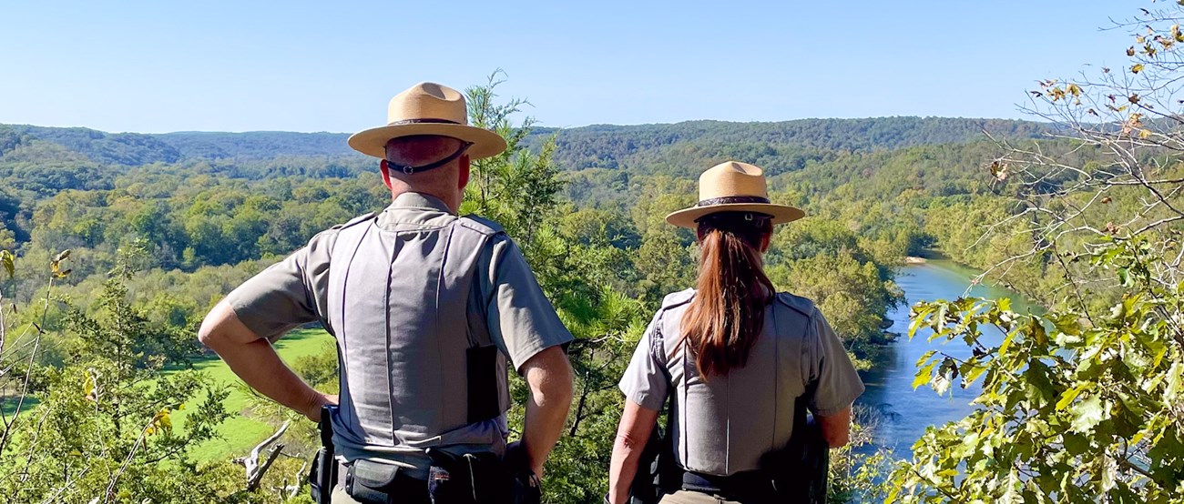 Two park rangers look down at a river and surrounding woods.