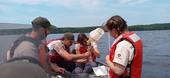 4 individuals sitting on a boat. 3 of the individuals are filtering water through a plankton net.