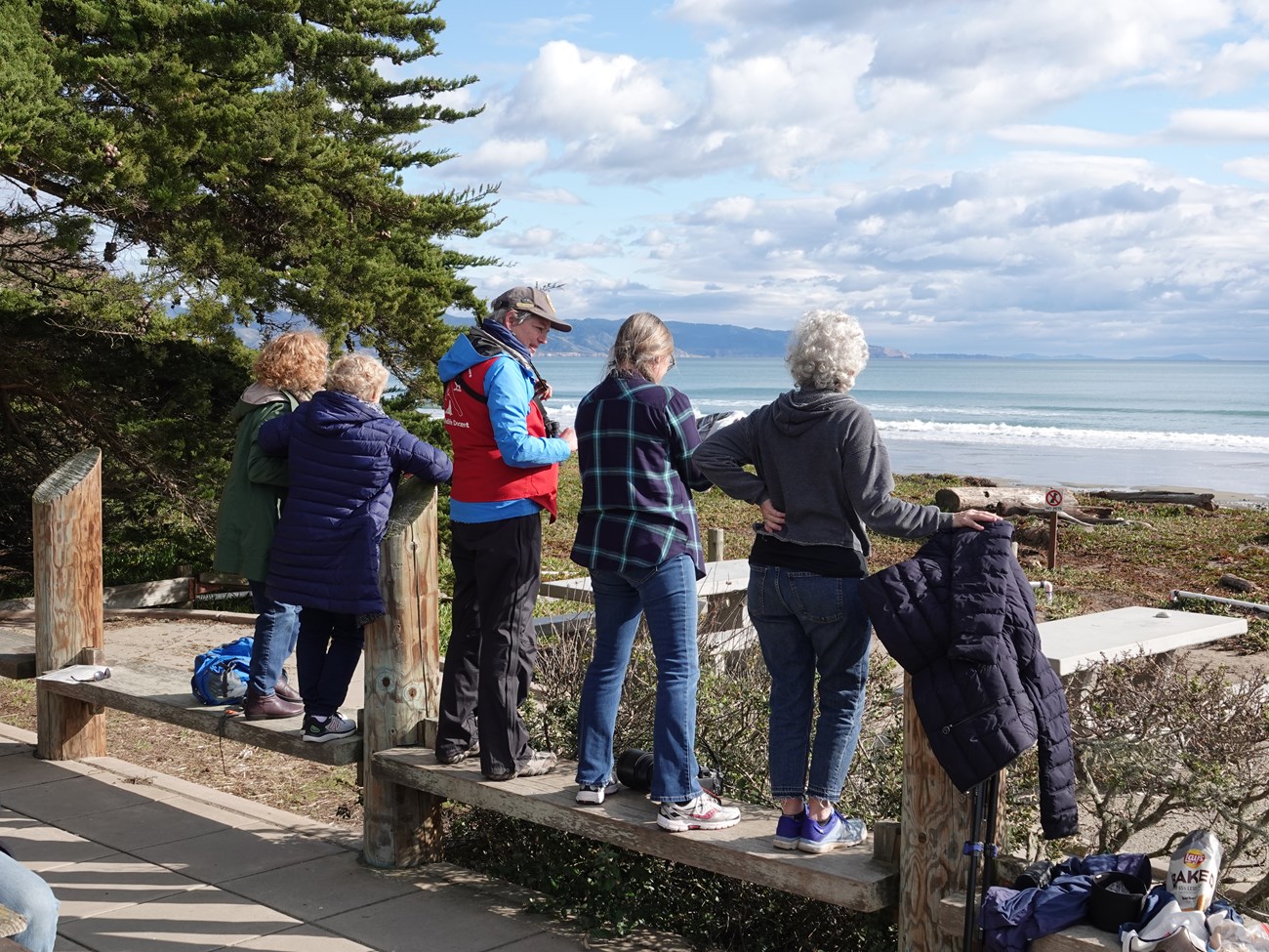 A volunteer in a red vest standing on a bench talks to park visitors near a beach.