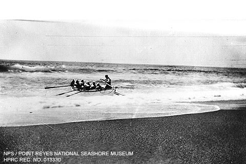 A black and white photo of seven men in a large rowboat rowing away from a beach through moderate surf.