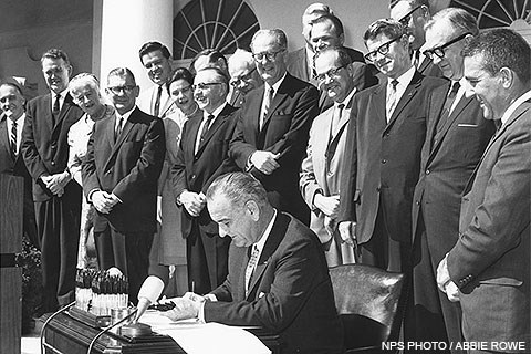 President Lyndon B. Johnson signs the Wilderness Act while fifteen men and two women stand behind him.