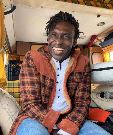 A black man wearing a plaid jacket laughs while sitting in a small camper van.