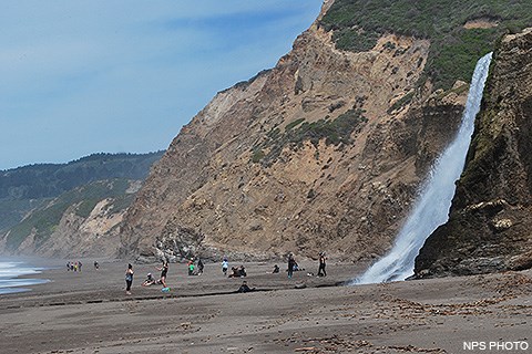 Twenty visitors playing and resting on an ocean beach. Pacific Ocean waves wash in from the left and a waterfall cascades onto the beach from coast bluffs on the right.