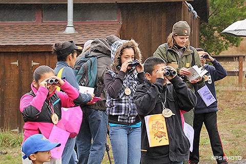 A park ranger consults a bird book as children around her look through binoculars.