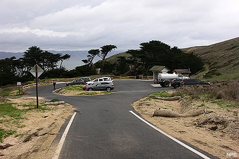 Ten vehicles parked in a small, parking lot. A vault toilet and a bus shelter are on the right side. Trees and the ocean are visible in the background.