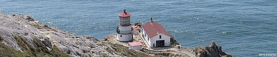 On the edge of the Pacific Ocean, a white, three-story tower with a red roof stands adjacent to three other red-roofed, white-sided buildings on. Barren rocks slope down to the lighthouse on the left.