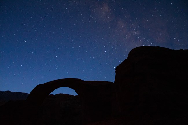 The Milky Way stretches across a dark blue night sky with the blackened silhouette of a natural stone bridge in the foreground.