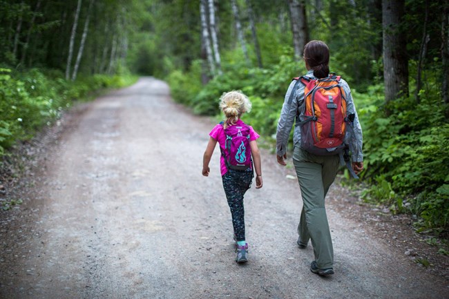 A little girl and a woman scientist step in sync down a gravel road in the woods.