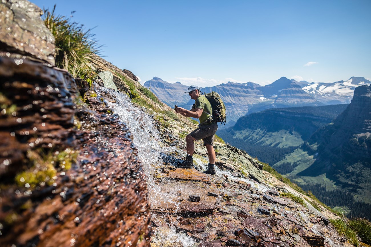 Man stands near a stream of water that juts out of a mountainside.