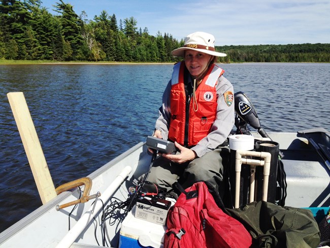 A woman wearing a National Park Service uniform and life jacket sits in a boat on a lake and holds a piece of monitoring equipment.