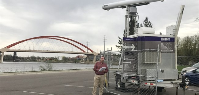 A special trailer with avian radar monitoring equipment is set up behind a fence. The Clark Bridge appears in the background.