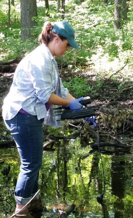 A woman standing in shallow water is holding a small electronic device attached to a long cord which dangles into the water.