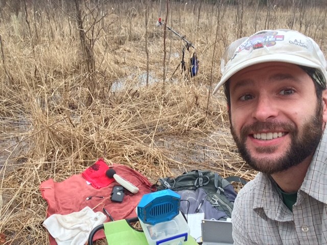 A man in a wetland smiles at the camera. His equipment, including microphones and speakers, is visible in the background.