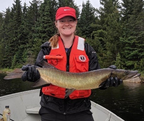 A woman in the bow of a boat smiles.  She is wearing a life jacket and rubber gloves, and is holding a large fish in her hands.