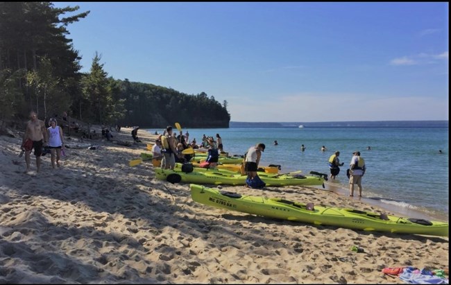 People walk along the beach past large numbers of yellow sea kayaks that have pulled up onto the shore.
