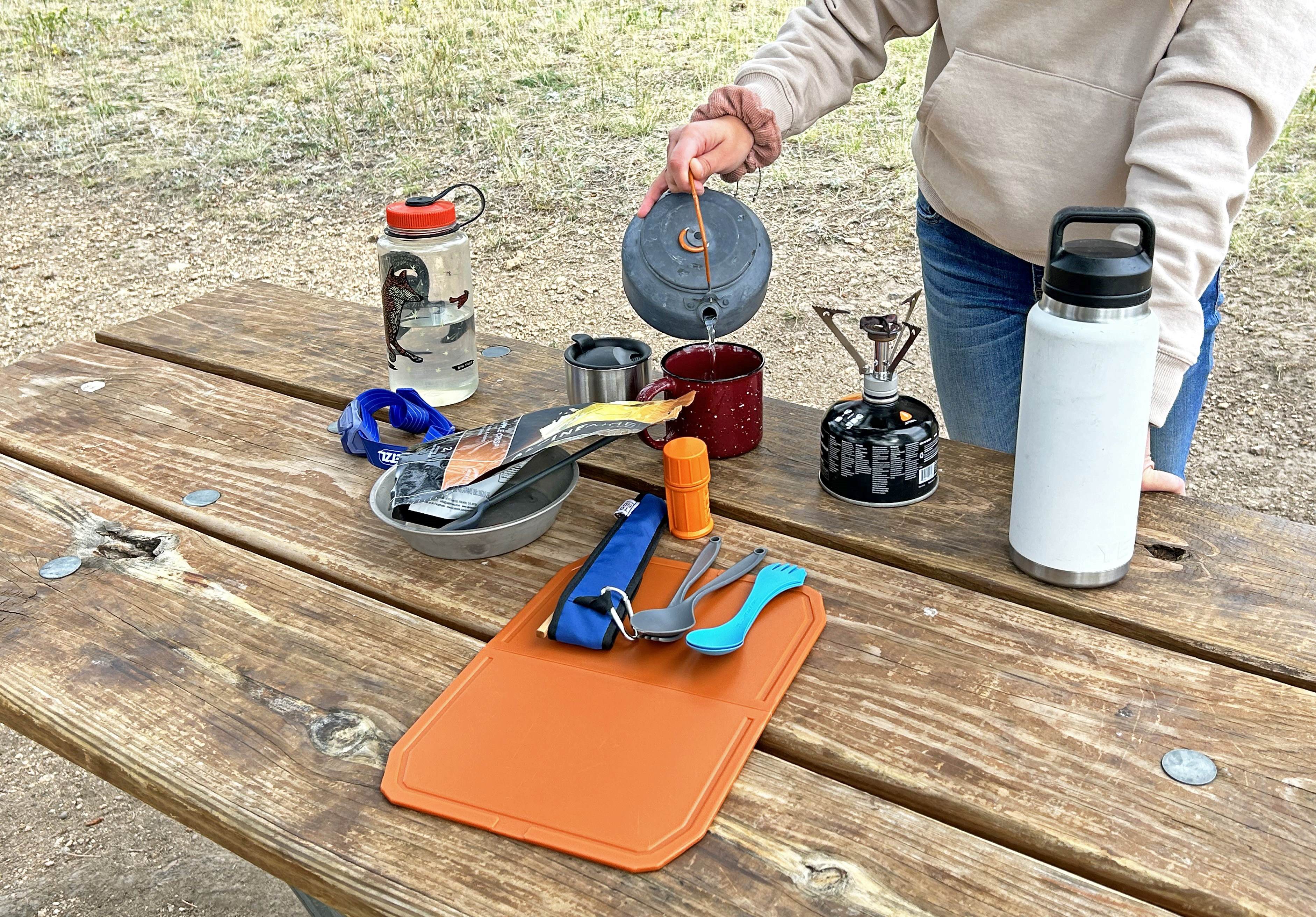 Camper is pouring hot water into a cup next to camp stove on top of a picnic table..