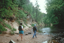Two people stand on a pebbly beach with a sandstone white rock face with some vegetation