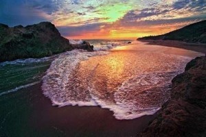 Image of Leo Carrillo State Park. Distant colorful clouds are back-lit by a sunset. It is framed by a rocky outcropping on the left and mountain slopes on the right. In the foreground: the Pacific Ocean waves with white, yellow, orange, and pink highlights gently slide up a smooth sandy beach.