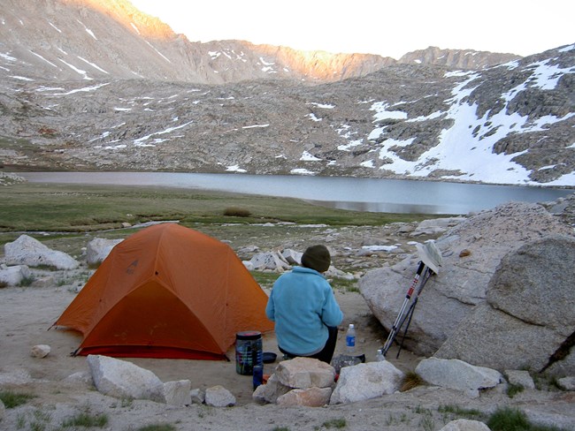 A camper sitting by a tent looking out at a lake and snowy mountains.